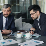 Two male business executives reviewing financial data and charts on a laptop during an investment consulting meeting in an office setting.
