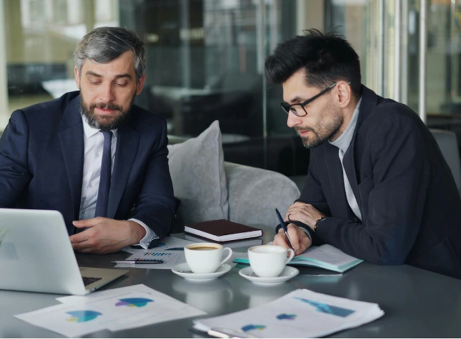 Two male business executives reviewing financial data and charts on a laptop during an investment consulting meeting in an office setting.
